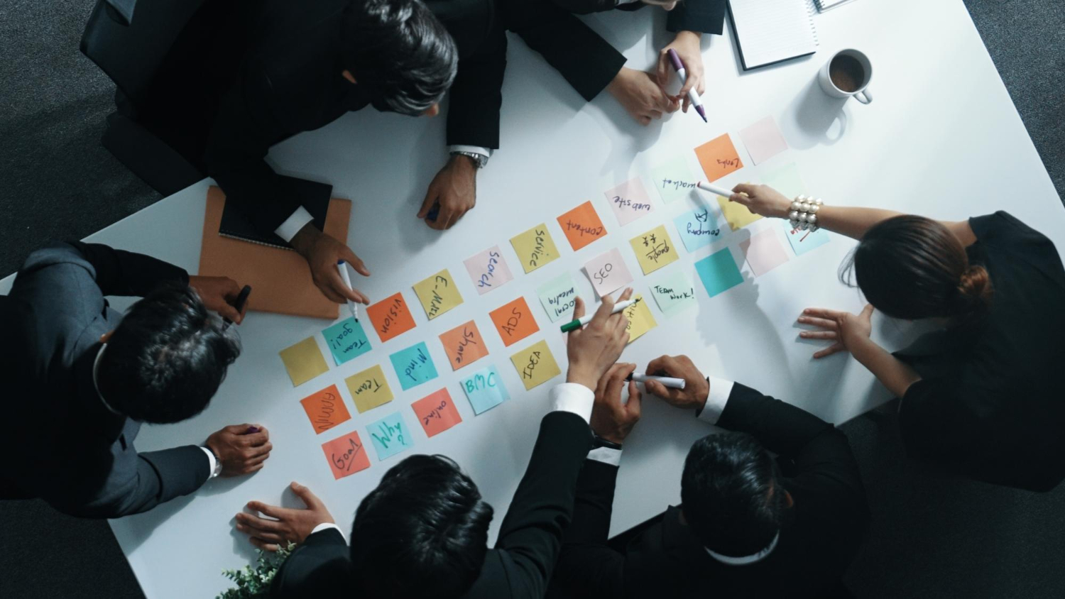 "Várias pessoas em trajes formais, sentadas em torno de uma mesa de reunião, organizando post-its coloridos com anotações estratégicas de negócios. A imagem mostra uma abordagem colaborativa de planejamento e brainstorming empresarial.